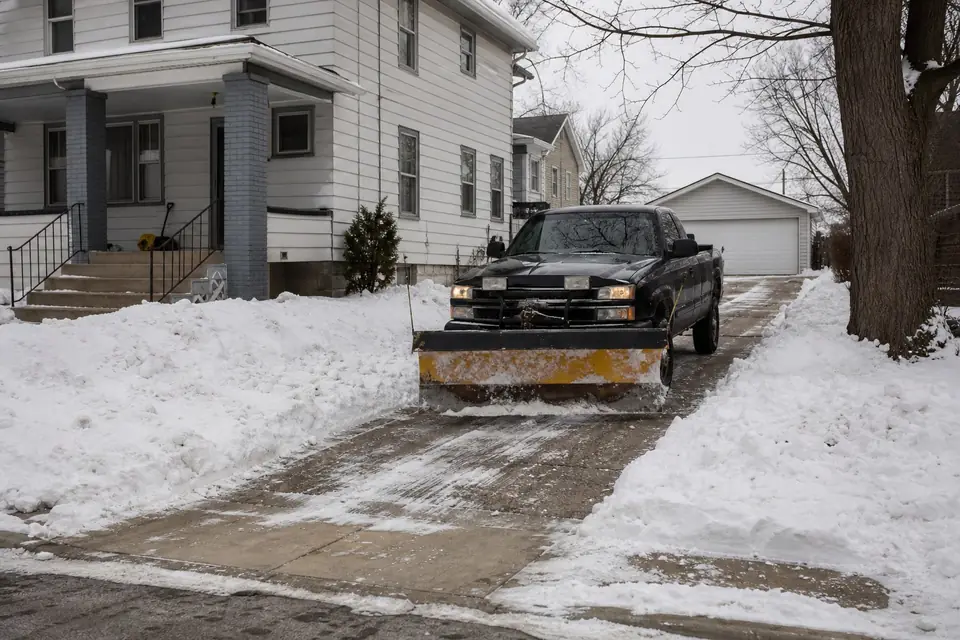 Plow truck clearing a long driveway beside an older house